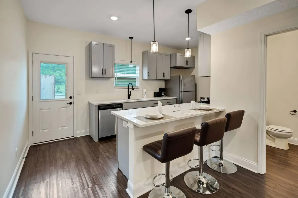 A kitchen scene with a sunlit island, bar stools, shiny appliances, and a large window. The space radiates warmth and sociability, illustrating a kitchen where hosting and everyday living intersect seamlessly.