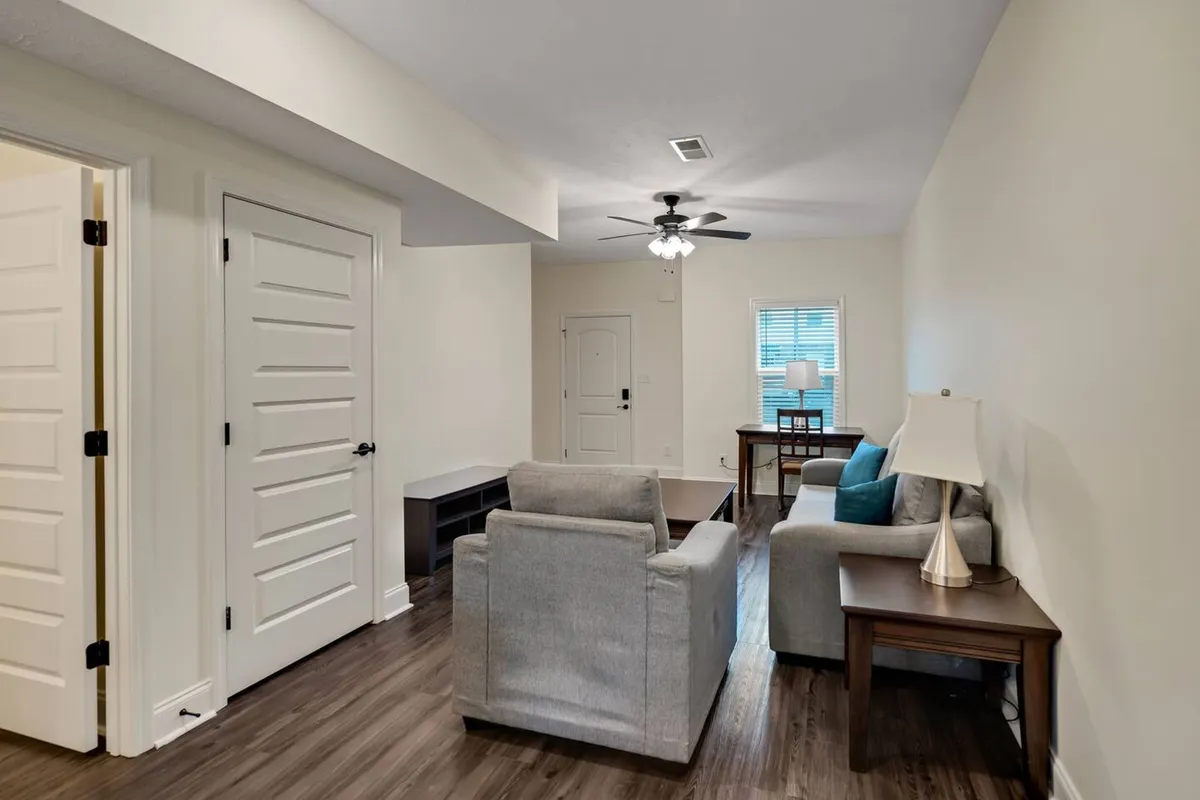 A bright living space featuring a sofa, a compact desk with a lamp, and a doorway allowing natural light to stream in. The setup is ideal for both unwinding and catching up on work, blending leisure with productivity seamlessly within the communal area.