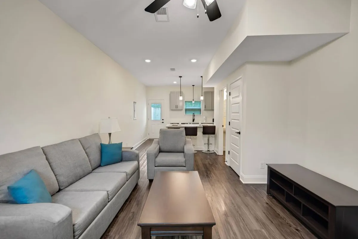 A stylish, sunlit living area with a plush sofa, inviting armchair, modern coffee table, and open kitchen—perfect for relaxing or gathering with friends. The space feels cozy, contemporary, and designed for both comfort and connection.
