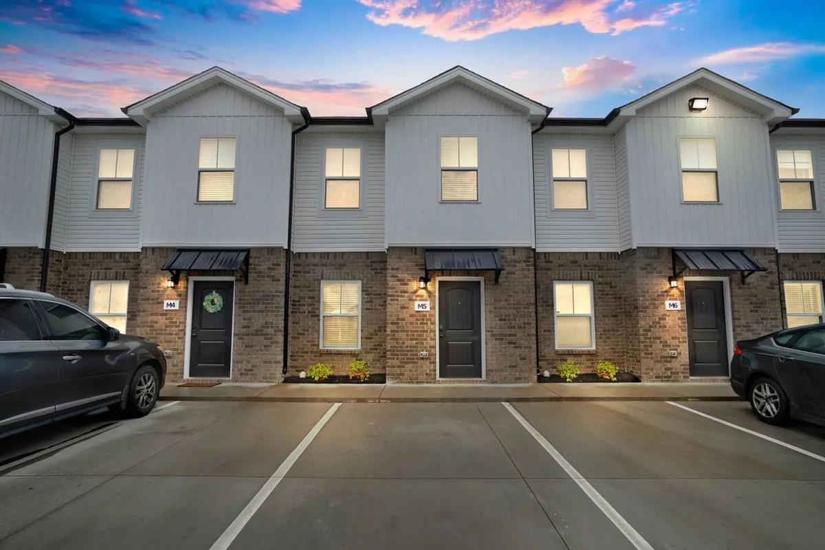 A wide view of the townhouse facades with sunset skies, decorative wreaths on doors, and a visible parking area. The image captures the entrance and easy vehicle access, creating a sense of both community and practicality for those coming and going for work.