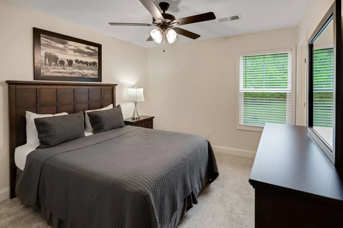 A sunlit bedroom with a plush bed, lamp, framed artwork, dresser, and windows that allow natural light to pour in. The space feels cozy and serene, evoking a sense of waking up refreshed and enjoying peaceful sleep.