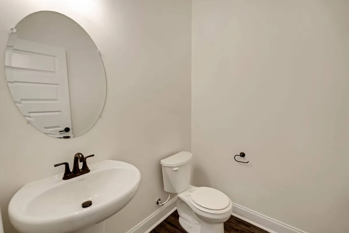 A serene bathroom featuring an oval mirror above the sink, with a toilet beside it. The space is bathed in soft, natural light, evoking a calm and simplistic mood. Minimal décor and clean lines enhance the sense of tranquility and refreshment.